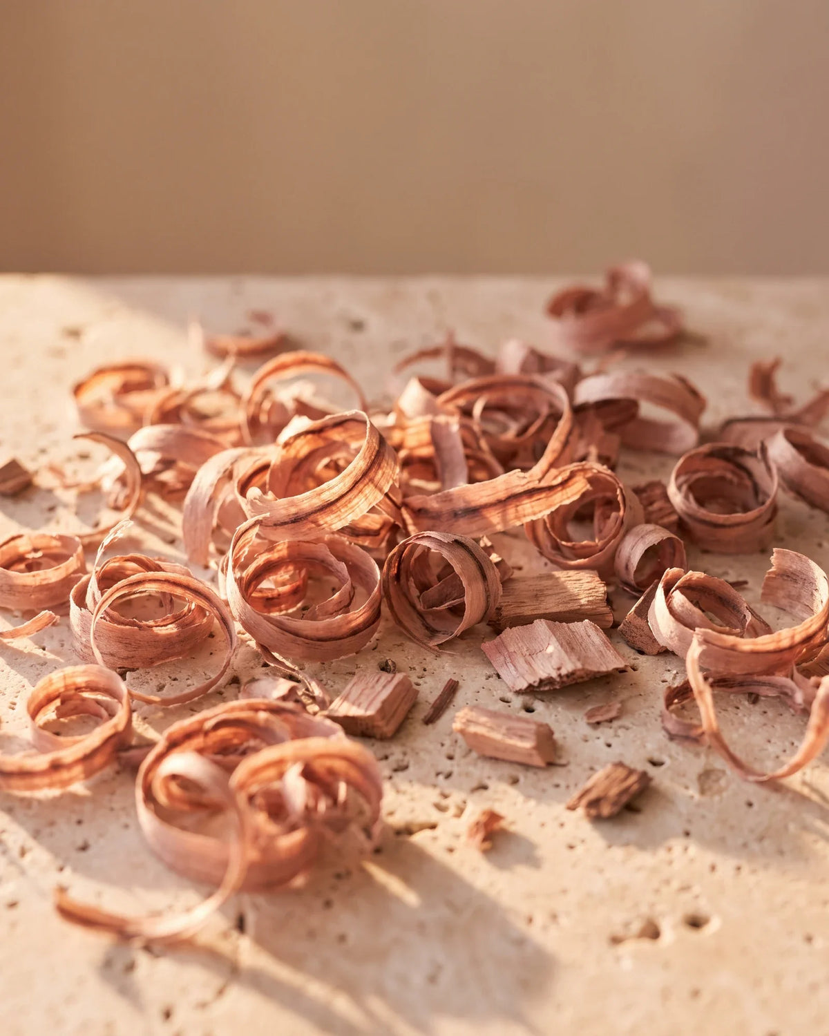 Finas lascas rosadas de pau-rosa brilhando sob luz quente sobre travertino — o aroma doce, floral e amadeirado do Óleo essencial puro de pau-rosa brasileiro Delune