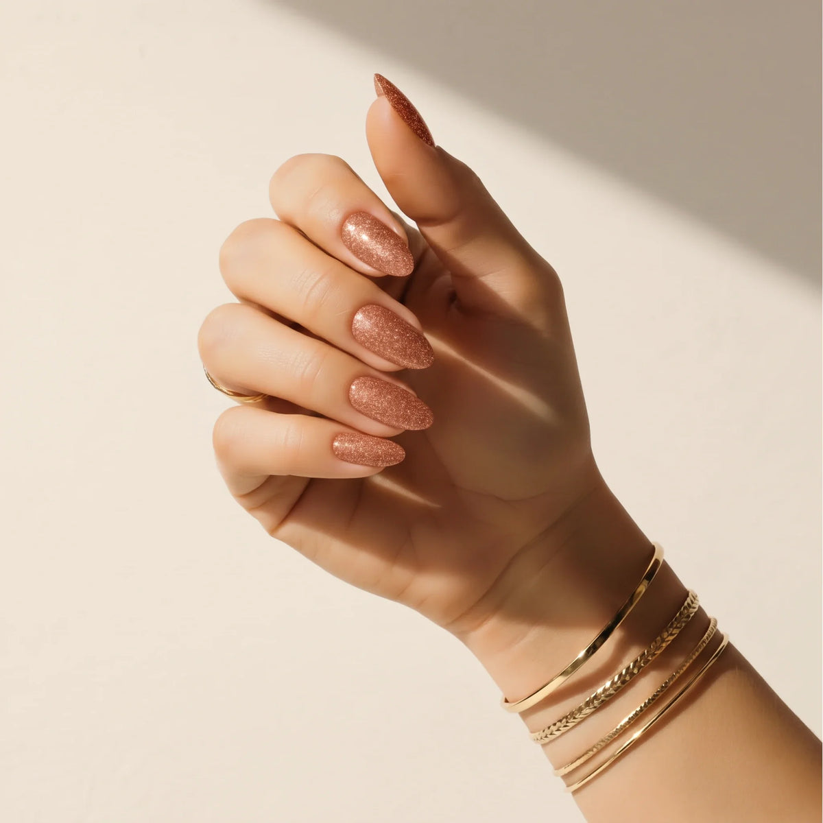 Mão com adesivos de unha Copper Glitter Delune e braceletes dourados contra uma parede de reboco quente