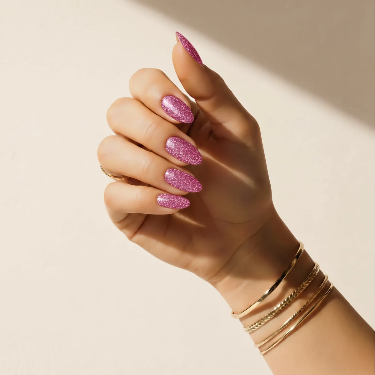 Mão com adesivos de unha Glitter Bug com glitter rosa Delune e braceletes dourados contra uma parede de reboco quente