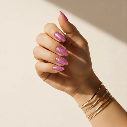 Mão com adesivos de unha Glitter Bug com glitter rosa Delune e braceletes dourados contra uma parede de reboco quente
