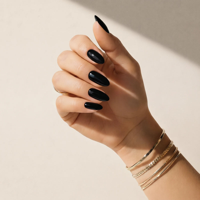 Mão com adesivos de unha Noir pretos brilhantes Delune e braceletes dourados contra uma parede de reboco quente